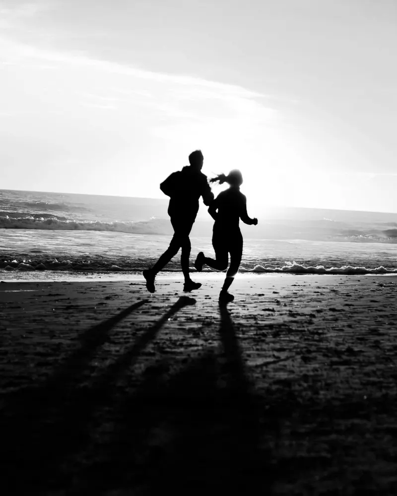 Two people running side by side along a beach at sunset, in black and white.
